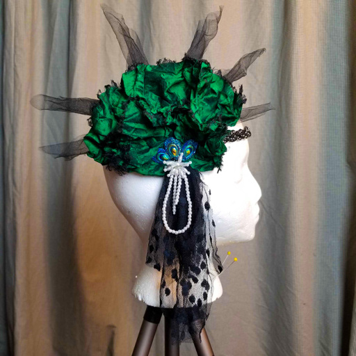 A flapper-esque headpiece with a beaded band across the forehead supporting a large green silk rosette over the ear with six black tulle spikes radiating from the top, and a fall of elegant dot mesh below. There are three beads resembling peacock feathers in the center of the silk rosette, above a pearl starbust with pearl strands contrasting against the dot mesh.