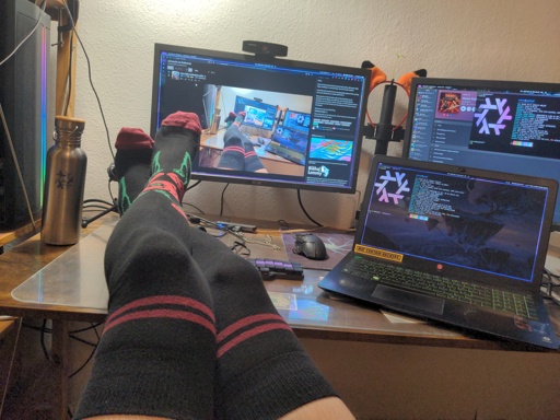 Typical unixsocks frame: Desk with socked legs on it. Black socks with red flowers in the ankle region and other red accents. Left, barely in the frame is a desktop computer with rainbow led stripes. On the desk are 2 screens from the desktop PC and laptop. Fuck there are so many details, I could mention. If you want an extended image description ask me in the comments.