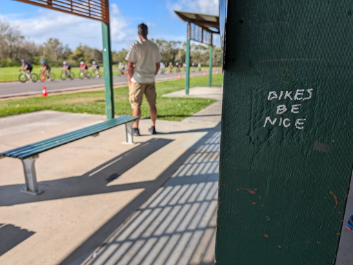 "Bikes be nice" written on the side of a post holding up the shade cover over a seated area next to a bike track. In the background, out of focus, a man watches a group of bikes racing past.
