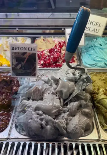 Image of ice-cream buckets in ice cream shop. In the center theres a gray ice-cream (cement color) and a constructiom trowel stuck in instead of a scoop. Theres also a small photo on a plastic stick inside the bucket (as a description label) showing a bucket of cement saying BETON EIS.