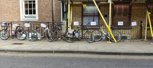 A fence with a4 paper with "no bikes" and "no signs" ziptied to them. There are 5 bikes lent against the fence
